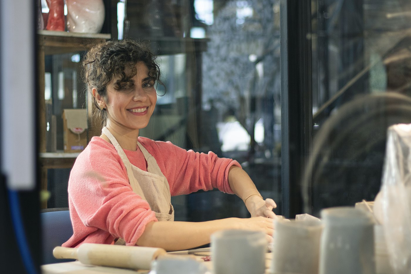 A smiling potter in an apron at a workbench full of clay pots